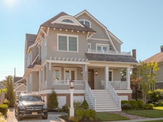 Light neutral colored tan siding with white trim, square columns and railings. Red brick veneer. Brown stained front door. Brown shingled roof. Beautiful beach house design.