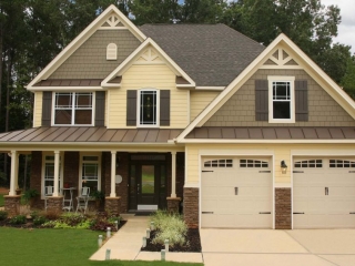 Two toned gray and pale yellow siding color combination. White trim and columns. Black roofing shingles with a brown metal accent roofline. White garage doors. Dark brown stained front door. Brown stone veneer. Gray shutters.