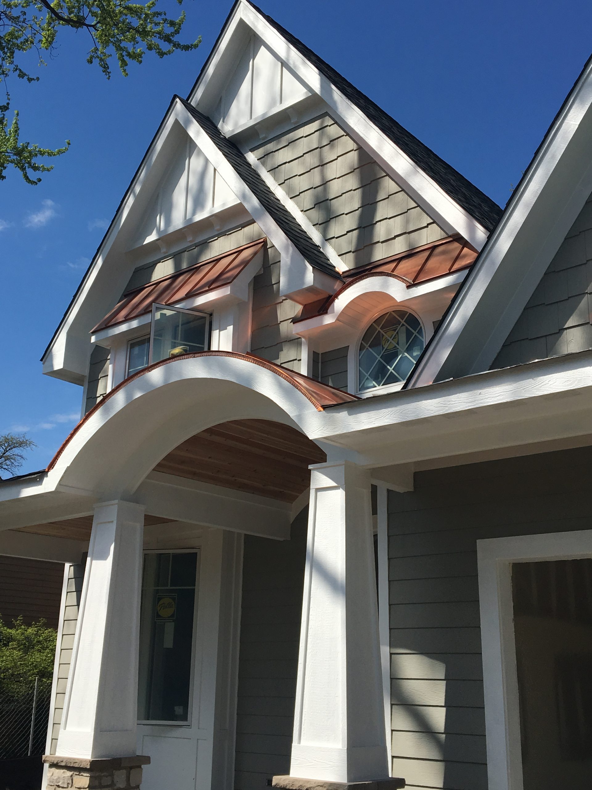 Gray siding colors with white trim. Brown metal roofing. Arched porch with tapered columns. Brown stone veneer.