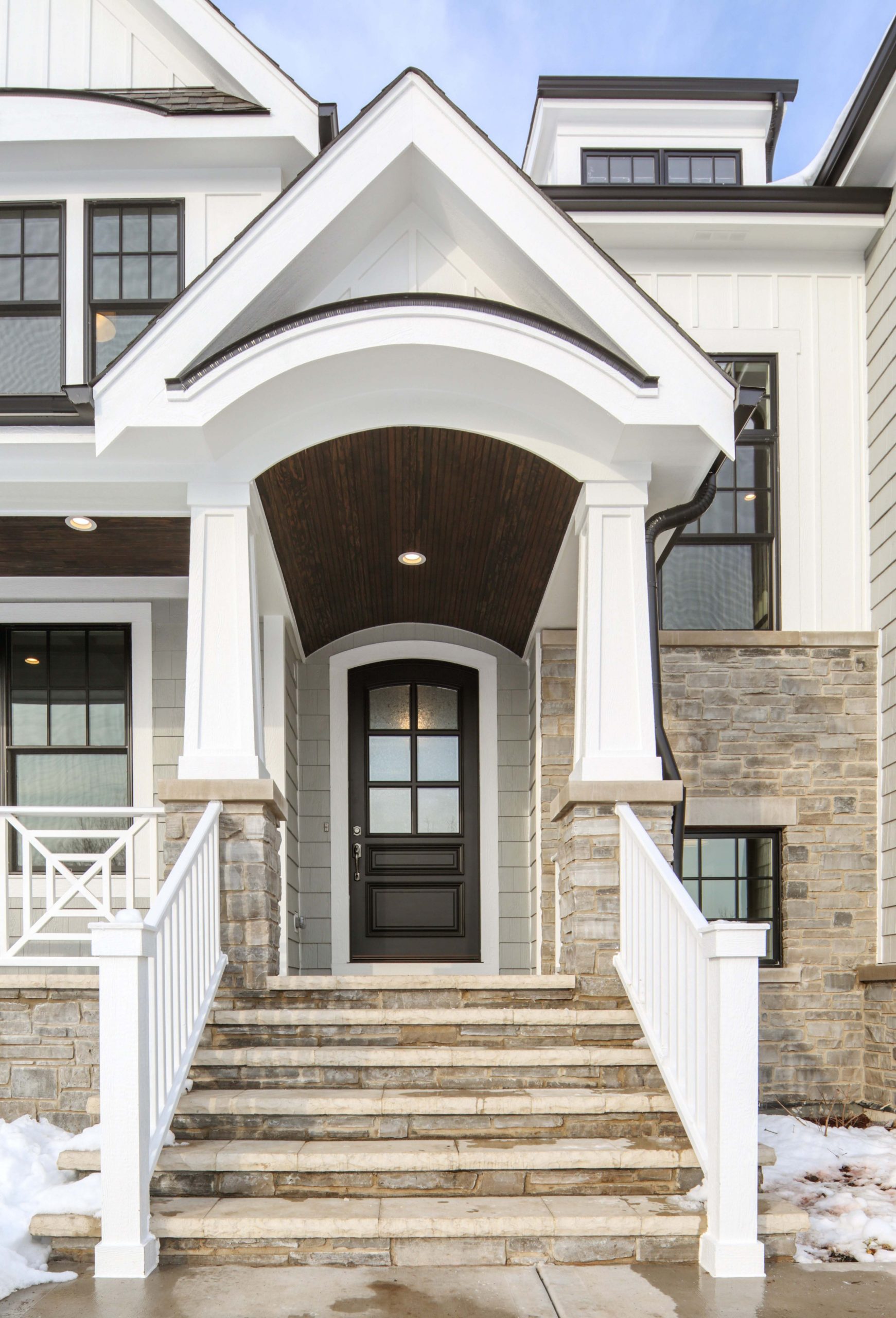 Light gray siding color with tons of white trim and white wall paneling. Dark brown wood porch soffit with recessed lighting. Tapered porch columns with white rails. Stone veneer. Black gutters. Black framed windows.