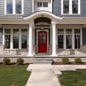 blue vinyl siding color with white trim, wall paneling and round columns. Red front door. Dark brown wood porch soffit. Light brown stone veneer. Concrete walkways.