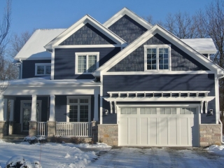 Dark blue colored siding with white trim. White tapered porch columns with white railings. Natural light brown stone veneer. White gutters. White garage doors.