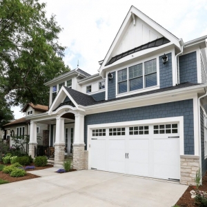 Blue vinyl siding colors. Light tan stone veneer. White garage door. White trim and wall paneling with blakc metal accent roof. Black shingled roof. Tapered porch columns. Black porch railings.