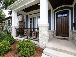 Blue siding color with brown stained front door. Black porch railings with white tapered porch columns. Concrete porch and steps. Light stone veneer. Dark brown stained wood porch soffit with recessed lighting.