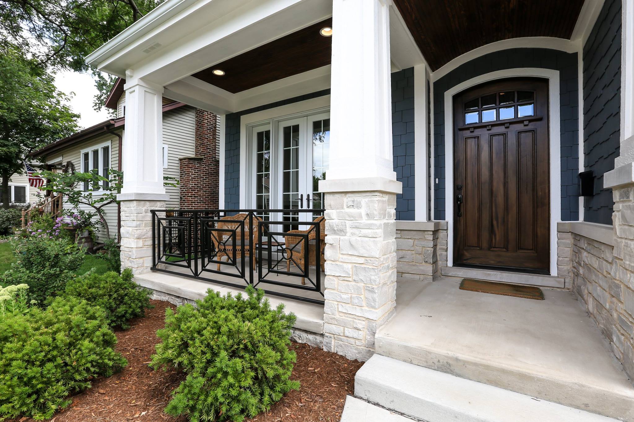 Blue siding color with brown stained front door. Black porch railings with white tapered porch columns. Concrete porch and steps. Light stone veneer. Dark brown stained wood porch soffit with recessed lighting.