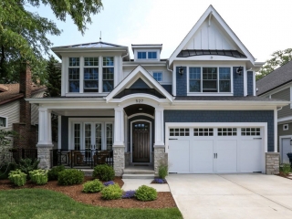 Blue cedar shake siding colors with white trim and wall paneling. Black shingled roof with metal accents. Dark brown stained wood front door with white garage doors. Light stone veneer. Dark wood stained porch soffit with recessed lighting. Concrete driveway.