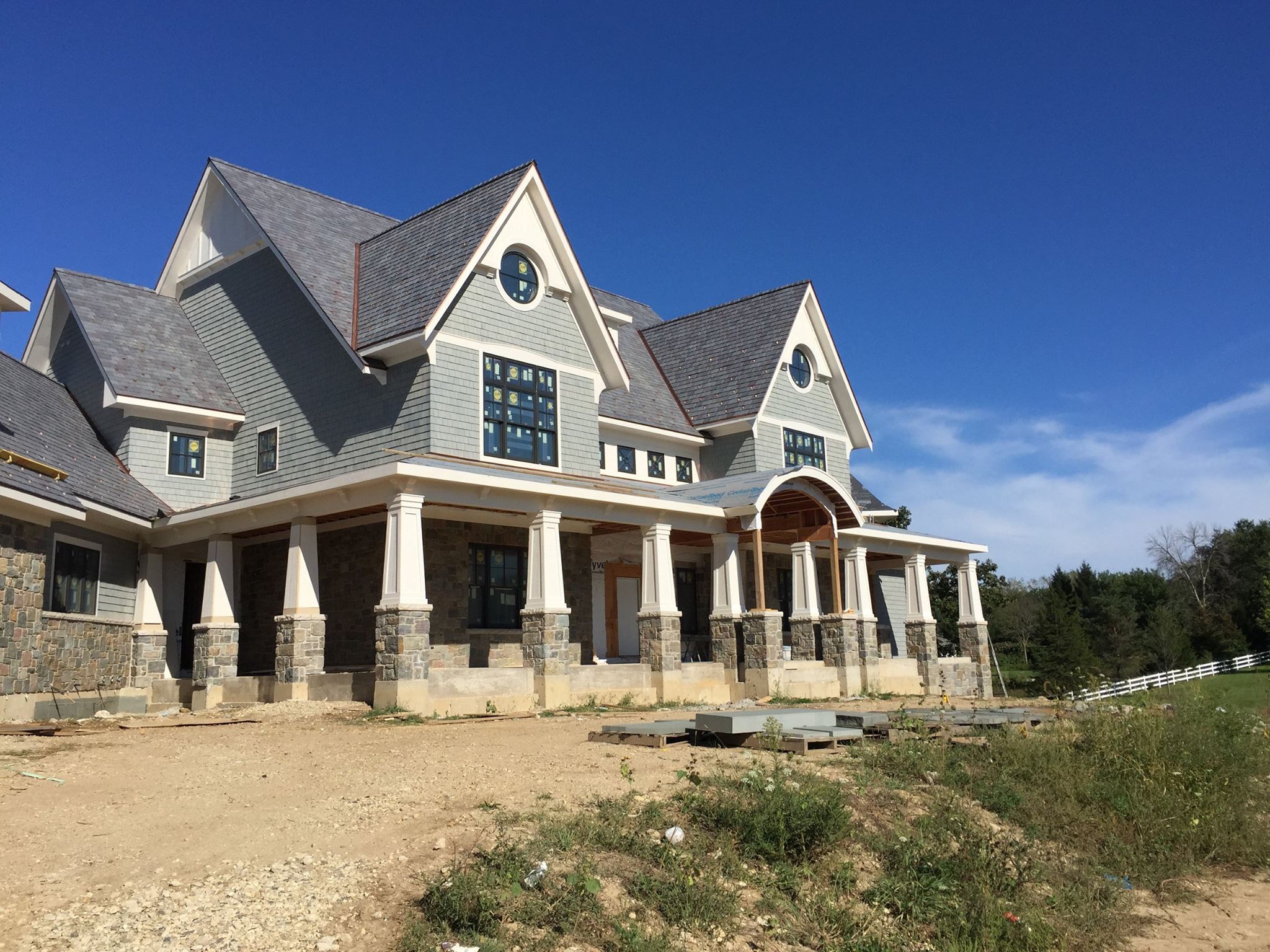 Light gray cedar shake siding with white trim and wall paneling. Tapered porch columns with stone veneer bases. Black framed windows. Brown stone veneer.