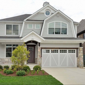Light gray siding colors with white trim and wall paneling. Dark brown front door. Light brown stone veneer. Concrete driveway and porch. White railings. White and gray garage doors. Dark brown wood stained porch soffit. Black shingled roof.