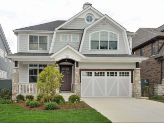 Light gray siding colors with white trim and wall paneling. Dark brown front door. Light brown stone veneer. Concrete driveway and porch. White railings. White and gray garage doors. Dark brown wood stained porch soffit. Black shingled roof.