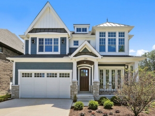 Blue vinyl siding colors with white trim and wall paneling, black metal roofing, brown stained wood front door, white tapered columns, light brown stone veneer, black railings, concrete driveway and walk.