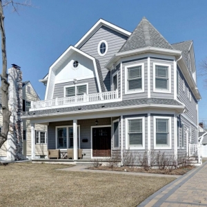 Gray viny siding color with white trim and wall paneling. Dark brown stained wood front door. White railings. Gray shingled roof. Paver driveway. Gray decking.