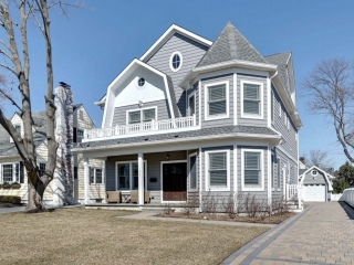 Gray viny siding color with white trim and wall paneling. Dark brown stained wood front door. White railings. Gray shingled roof. Paver driveway. Gray decking.