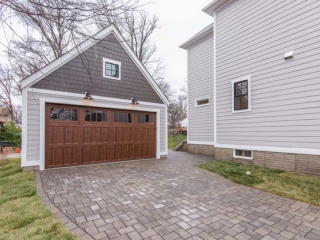 Two car detached garage design with one large door. Two toned gray siding with a stained brown door. Paver driveway. Black framed windows.