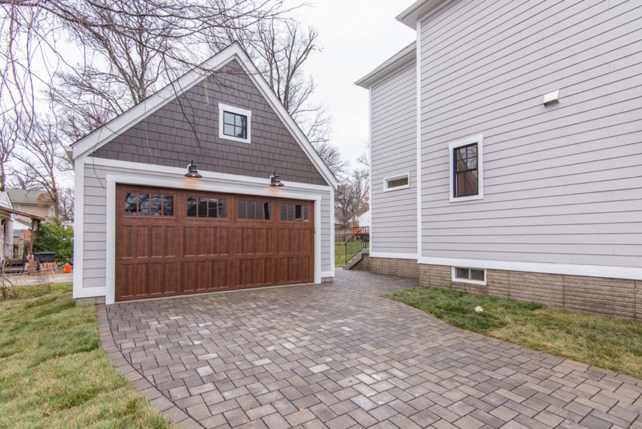 Two car detached garage design with one large door. Two toned gray siding with a stained brown door. Paver driveway. Black framed windows.