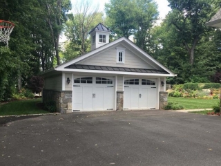 2 Car detached garage design. Light tan siding with white garage doors and real stone veneer. Black metal accent roofing.