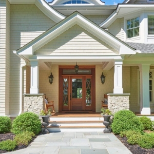 Neutral house siding colors. Light tan with a brown stained wood front door. Light gray stone veneer. White trim. White columns. Blue stone walkway.