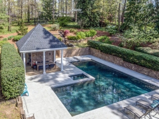 Small pool house design with red brick walls and a black shingle roof. Concrete patio. Rectangle in ground pool. Covered seating area. Red brick retaining walls. Poolside seating.