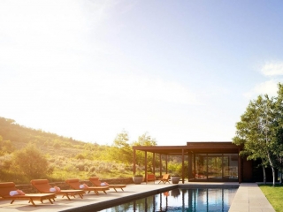 Beautiful modern pool house design in the country at sunrise. Huge floor to ceiling windows. Metal and wood siding. In ground rectangle pool. Poolside seating. Stone retaining walls. Flat roof.
