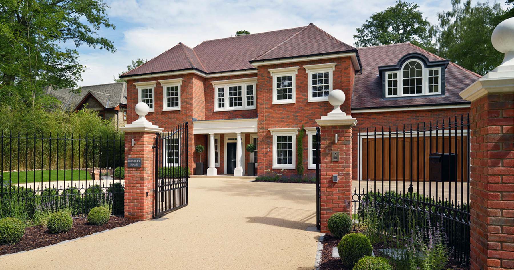 Beautiful custom red brick home with purple gray roof shingle. Black front door. Tan stone trim and columns. Arched dormers. Red brick pillars with black gate and stone cap.