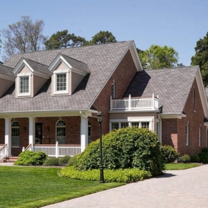 Red brick ranch home design. Gray brown roof color with dormers. White columns, trim and railings with wood hand rail. White garage doors.