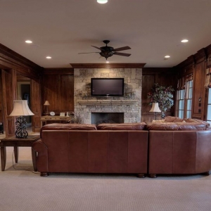 Living room with all wood walls, beige carpet and white ceiling. Brown leather sofa. Stone fireplace.