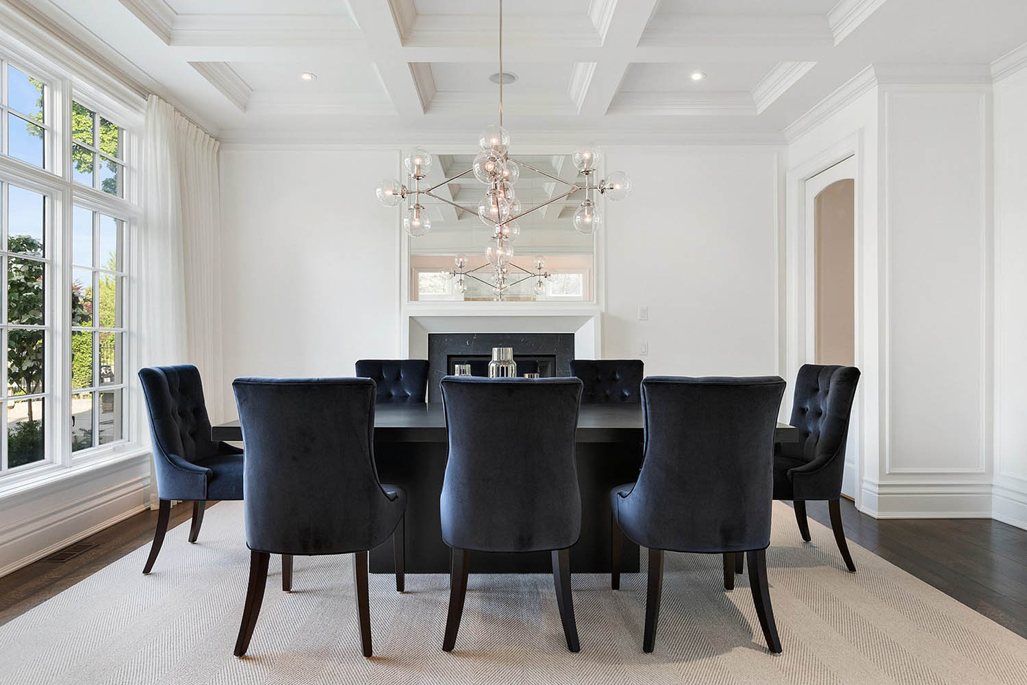 All white dining room with white coffered ceiling with black table and plush chairs. Dark hardwood floors with tan area rug.