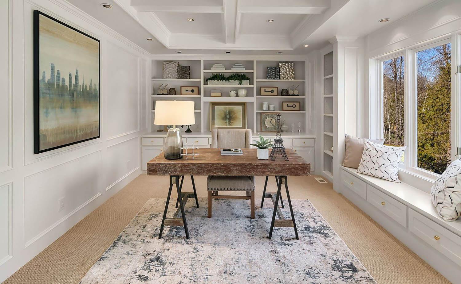 Home office coffered ceiling with soffits. All white ceiling and walls with white trim and wall paneling. Wood desk with metal black legs. Built ins and window seat.
