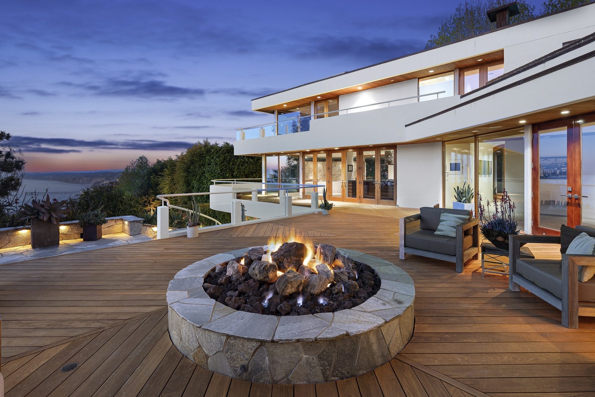 Modern home with white stucco siding mixed with light wood frames and soffits. Custom wood deck with stone fire pit.