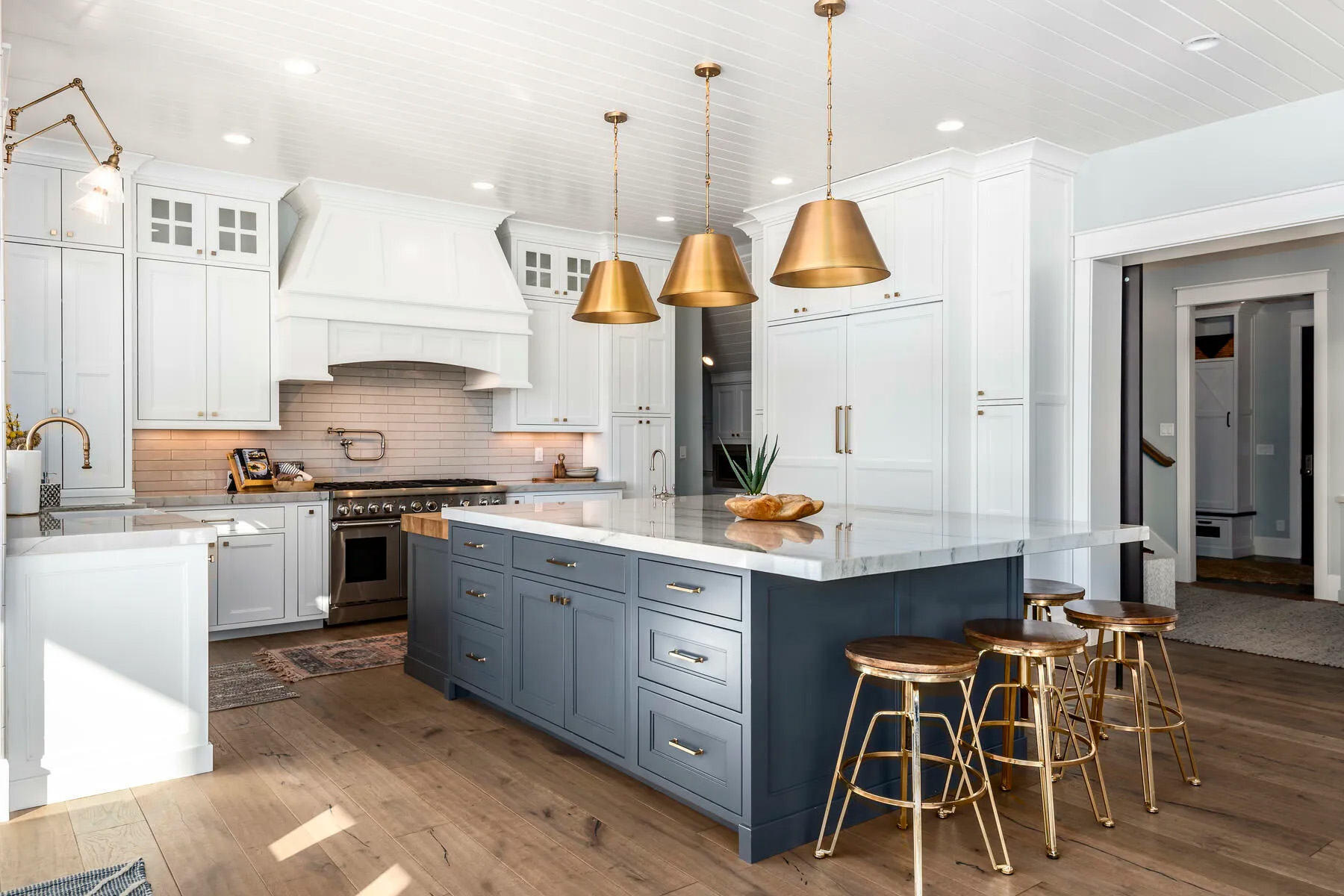 Stunning kitchen design including a blue island, marble countertops, white wall cabinetry and gold and wooden stools.